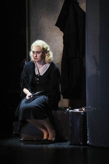 Vaaleatukkainen nainen, jolla on musta huivi ja mekko, istuu matkalaukun päällä hämärässä huoneessa ja näyttää mietteliäältä. Takki roikkuu seinällä hänen takanaan, ja toinen matkalaukku on lähellä.