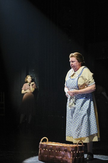 Kuviolliseen mekkoon ja esiliinaan pukeutunut nainen seisoo valokeilassa kangas kädessään ja kori jaloissaan. Hämärässä taustalla toinen nainen nojaa seinää vasten, osittain varjossa.