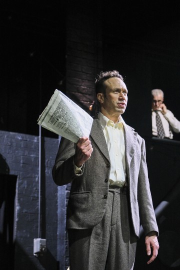 Harmaapukuinen mies, jolla on kädessään sanomalehti, näyttää huolestuneelta näyttämöllä dramaattisessa valaistuksessa. Taustalla toinen mies seisoo korkeammalla ja puhuu puhelimeen.