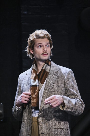 Nuori mies, jolla on vaaleat, aaltoilevat hiukset, harmaa kuvioitu bleiseri ja kuviollinen huivi, seisoo tummalla taustalla ja katsoo hieman sivulle yllättyneenä tai uteliaana.