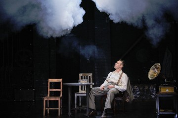 Vintage-vaatteisiin pukeutunut mies istuu pöydän ääressä hämärästi valaistulla näyttämöllä ja katselee ylöspäin yläpuolellaan olevaa savupilveä. Hänen vieressään on vanha gramofoni ja tyhjiä tuoleja, jotka luovat dramaattisen ja nostalgisen tunnelman.