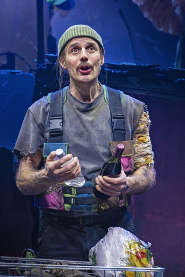 Tilkkutäkkipaitaan ja vihreään pipoon pukeutunut henkilö seisoo sisätiloissa, kädessään viinipullo ja muovipullo, edessään ostoskärryt täynnä tavaroita, ja katsoo ilmeikkäästi ylöspäin.