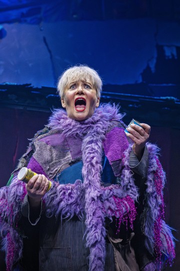 Vanhempi nainen, jolla on lyhyet harmaat hiukset ja jolla on yllään värikäs tilkkutäkki ja karvainen violetti huivi, pitää kahta pilleripulloa ja laulaa intohimoisesti lavalla dramaattisessa valaistuksessa.