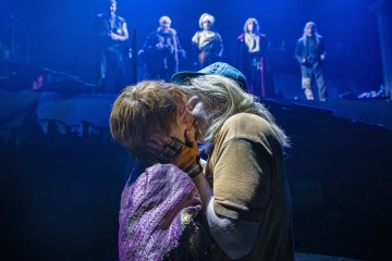 Kaksi ihmistä syleilee ja suutelee intohimoisesti dramaattisen sinisen näyttämövalaistuksen alla, kun viisi muuta ihmistä seisoo taustalla korotetulla korokkeella ja katselee kohtauksen kulkua.