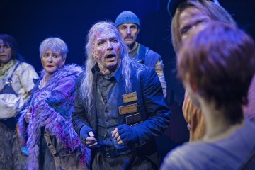 Rähjäisiin, värikkäisiin pukuihin pukeutuneet näyttelijät seisovat näyttämöllä dramaattisessa valaistuksessa, heidän kasvonsa ovat ilmeikkäät ja likaiset, mikä viittaa teatteriesityksen intensiiviseen hetkeen.