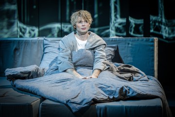 Nuori henkilö, jolla on vaaleat hiukset, istuu pystyasennossa sängyssä, harmaaseen peittoon käärittynä, vaalea kaapu yllään ja mietteliään näköisenä. Huone on hämärästi valaistu ja siinä on viileän sinisiä sävyjä ja abstrakti tausta.