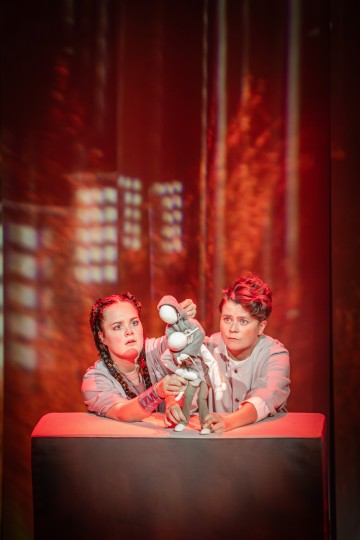 Kaksi esiintyjää, joilla on vakavat ilmeet, manipuloivat harmaata humanoidinukkea pöydällä, taustalla on dramaattinen punainen valaistus ja abstrakteja kuvioita, jotka luovat teatraalisen ja intensiivisen kohtauksen.