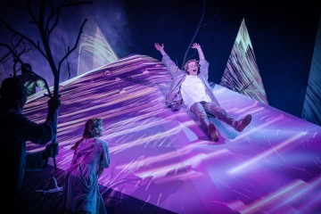 Vaaleisiin vaatteisiin pukeutunut henkilö liukuu alas hehkuvaa, vinoa pintaa, jossa on violetteja ja valkoisia raitoja, kun muut katselevat lähistöllä abstraktien, vuorten kaltaisten rakenteiden ja sumun keskellä. Kohtaus on dramaattinen ja teatraalinen.