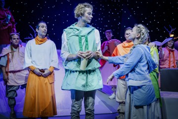 Ryhmä värikkäisiin pukuihin pukeutuneita näyttelijöitä esiintyy lavalla tähtitaivaan taustaa vasten; yksi vihreäpukuinen henkilö seisoo keskellä pitelemässä kääröä, kun toinen sinipukuinen henkilö pitelee hellästi hänen kättään.