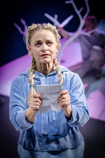 Vaaleat punotut hiukset omaava nainen, jolla on vaaleansininen huppari, pitää kädessään paperia ja katsoo huolestuneena ylöspäin. Taustalla näkyvät sumeat valkoiset puun oksat ja henkilö.