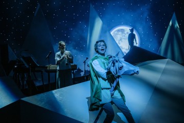 Vihreään viittaan pukeutunut esiintyjä pitelee valkoista hevosta dramaattisella, sinisellä valaistulla näyttämöllä, jossa on geometrisia muotoja, kun toinen henkilö seisoo mikrofonin takana. Taustalla näkyy täysikuu, jossa on siluettina hahmo.