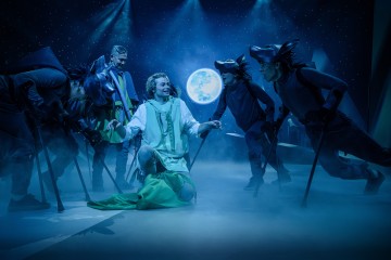 Näyttelijät esiintyvät sumuisella näyttämöllä tähtitaivaan ja täysikuun alla; yksi henkilö polvistuu valkoisissa ja vihreissä kaavuissa, ja ympärillä on muita, joilla on susipuvut, joissa on teräväkorvaiset ja maalatut kasvot.