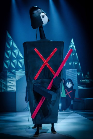 Lavalla seisoo esiintyjä, jolla on geometrinen musta puku, jossa on punainen X-kirjain ja suuret silmät, ja hänellä on kädessään punaisia tikkuja. Taustalla on abstrakteja, kulmikkaita maisemia ja toinen samanlaiseen asuun pukeutunut esiintyjä. Valaistus on dramaattinen ja sinisävyinen.