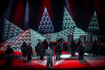 Tummiin hupullisiin kaapuihin ja punaisiin X-kirjaimiin pukeutuneet esiintyjät seisovat lavalla, jossa on geometrisia, pyramidimaisia rakenteita ja dramaattinen punainen valaistus, joka luo salaperäisen ja teatraalisen tunnelman.