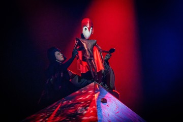 Mustiin pukeutunut esiintyjä ohjaa pyramidin päällä olevaa suurta, punapukuista nukkea, jolla on valkoiset, naamiomaiset kasvot, dramaattisen punaisen ja sinisen näyttämövalaistuksen alla.