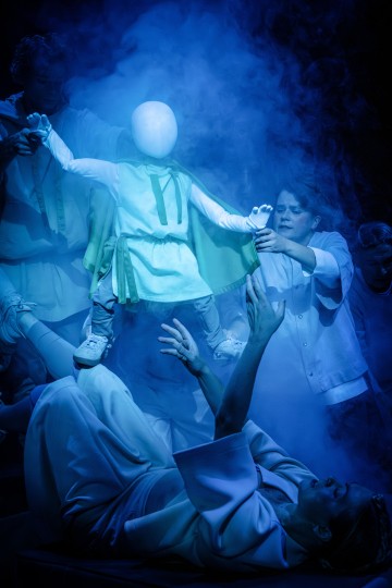 Useat ihmiset nostavat kasvotonta, valkoiseen pukuun pukeutunutta lapsimaista hahmoa pimeällä näyttämöllä pyörivän sinisen savun keskellä, mikä luo surrealistisen ja dramaattisen tunnelman.