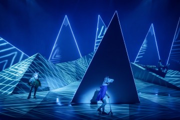 Kolme hevosnaamareihin pukeutunutta esiintyjää on lavalla suurten, valaistujen kolmionmuotoisten muotojen ja sinisten geometristen valokuvioiden ympäröimänä, mikä luo surrealistisen, futuristisen maiseman. Yksi esiintyjä ajaa skootterilla.