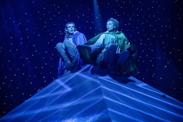 Kaksi värikkäisiin viittoihin pukeutunutta näyttelijää istuu näyttämöllä suuren, valaistun pyramidinmuotoisen rakennelman päällä, ja tähtitaivas korostaa dramaattista kohtausta.