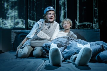 Kaksi näyttelijää istuu näyttämön sängyllä. Toinen, jolla on kypärä ja harmaat vaatteet, näyttää innostuneelta ja viittoo eteenpäin. Toinen makaa harmaan peiton alla, näyttää rennolta ja hymyilee. Tausta on tumma ja siinä on abstrakteja kuvioita.