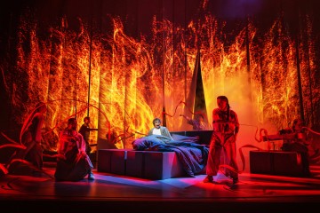 Dramaattisessa näyttämökohtauksessa keskushahmo makaa sängyllä, jota ympäröivät tanssijat, joilla on virtaavia nauhoja, eloisan, tulisen oranssin taustan edessä, mikä luo intensiivisen ja dynaamisen ilmapiirin.