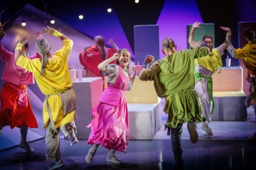 Joukko ihmisiä värikkäissä, yhteensovitetuissa asuissa tanssii iloisesti kirkkaasti valaistulla lavalla, jonka taustalla on geometrisia muotoja ja pastellivärisiä rekvisiittaa. Tunnelma on energinen ja vilkas.