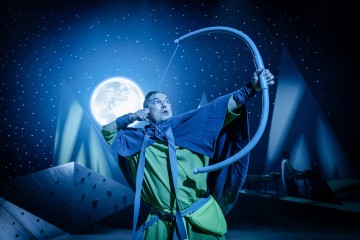 Vihreään ja siniseen pukuun pukeutunut henkilö vetää dramaattisesti suurta jousipyssyä ja tähtää nuolta kirkkaan täysikuun alla, ja taustalla näyttämöllä on geometrisia vuoria ja tähtitaivas.