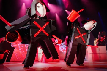 Kaksi esiintyjää, joilla on suuret mustat puvut, joissa on punaiset X-kirjaimet ja valkoiset, naamioituneet kasvot, pitelee punaisia rekvisiittaa kirkkaasti valaistulla näyttämöllä, jossa on vaaleanpunainen ja violetti tausta. Kohtaus vaikuttaa leikkisältä ja teatraaliselta.