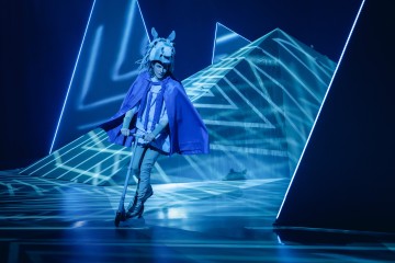 Henkilö, jolla on hevosnaamari ja sininen viitta, ajaa skootterilla lavalla, jonka taustalla on geometrinen sininen valaistus ja kuvioita.