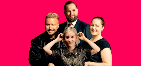 Neljä hymyilevää aikuista poseeraa yhdessä kirkkaan vaaleanpunaisen taustan edessä. Yksi edessä oleva nainen pitää leikkisästi sormiaan korvillaan, kun muut seisovat tiiviisti hänen takanaan.