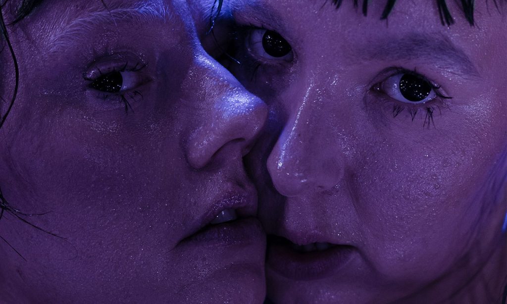 Kaksi ihmistä, joilla on märkä, kiiltävä iho, painaa kasvonsa tiiviisti yhteen dramaattisen violetin valaistuksen valaisemina, ja molemmat katsovat intensiivisesti kameraan.
