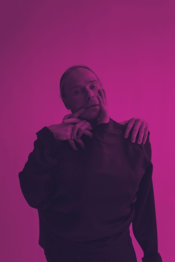 Henkilö seisoo vaaleanpunaista taustaa vasten toisen henkilön kädet kietoutuneina hänen ylävartalonsa ja kasvojensa ympärille, mikä luo surrealistisen, taiteellisen poseerauksen. Kuvassa on voimakas vaaleanpunainen värikerros.