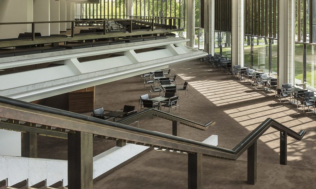 Tilava moderni sisustus, jossa on suuret ikkunat, ruskea matto, mustat pöydät ja tuolit sekä porrastetut parvekkeet. Auringonvalo virtaa korkeiden pystysuuntaisten ikkunalasien läpi ja luo varjoja lattiaan.