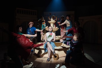 Nuori nainen istuu itsevarmasti suihkulähteen reunalla lavalla, ja ympärillä on värikkäisiin pukuihin pukeutuneita esiintyjiä, jotka poseeraavat dramaattisesti näyttämön valaistuksessa.