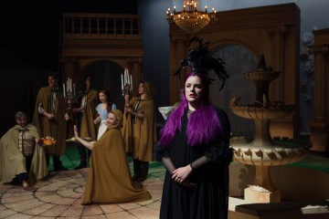 Etualalla seisoo nainen, jolla on pitkät purppuranpunaiset hiukset ja musta höyhenpäähine, ja ruskeisiin viittoihin pukeutuneet ihmiset ovat vuorovaikutuksessa suihkulähteen ja kattokruunun lähellä näyttämön kaltaisessa suuressa ympäristössä.