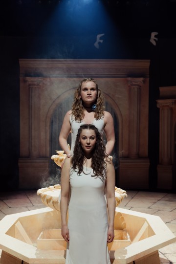 Kaksi valkoisiin mekkoihin pukeutunutta naista seisoo toistensa takana höyryävän suihkulähteen edessä, ja dramaattinen valaistus korostaa heitä klassisten kaarien taustaa vasten.