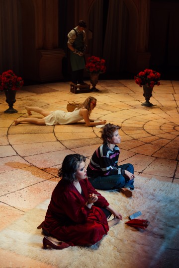 Etualalla vaalealla matolla istuu kaksi ihmistä, toinen punaisessa aamutakissa ja toinen rennoissa vaatteissa. Taustalla nainen makaa lattialla, ja toinen seisoo maljakoissa olevien kukkien vieressä, kaikki lämpimästi valaistulla näyttämöllä.