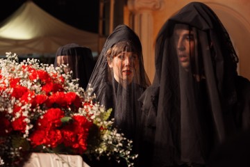 Kolme mustiin huntuihin pukeutunutta henkilöä seisoo juhlallisesti punavalkoisilla kukilla koristellun arkun vieressä, mikä viittaa hautajaisiin tai surukohtaukseen. Keskushenkilö katsoo eteenpäin synkkä ilme kasvoillaan.