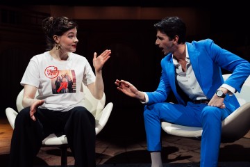 Valkoiseen T-paitaan painettu nainen ja kirkkaan siniseen pukuun pukeutunut mies istuvat valkoisilla tuoleilla ja käyvät vilkasta keskustelua hämärästi valaistulla näyttämöllä.