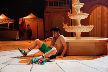 Vihreisiin shortseihin, lenkkareihin ja aurinkohattuun pukeutunut nuori mies istuu ilman paitaa ja väsyneen näköisenä suurta suihkulähdettä vasten kaakelilattialla. Taustalla näkyy telttoja, pöytä ja ihmisiä.