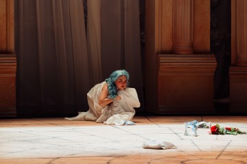 Sinihiuksinen, valkoiseen lakanaan kääritty nainen kyyristelee klassisten pylväiden muodostamalla näyttämöllä. Hänen lähellään lattialla on hajallaan ruusuja, mekko ja kenkiä. Kohtaus vaikuttaa dramaattiselta ja tunteikkaalta.