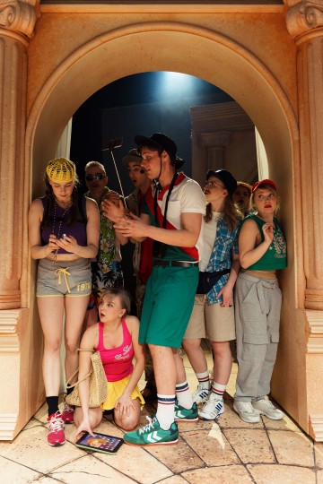 Ryhmä värikkäisiin, rentoihin vaatteisiin ja asusteisiin pukeutuneita nuoria kokoontuu koristeellisen kivikaaren alle, katselee elektronisia laitteita ja vaikuttaa uteliailta tai keskittyneiltä. Yksi henkilö polvistuu tabletin kanssa lattialle.