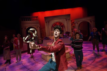 Nuori näyttelijä punaisessa takissa elehtii dramaattisesti keskellä näyttämöä, jota ympäröi värikäs puvustettu näyttelijäkaarti. Lavastuksessa on kaaria, pylväitä ja elävä punainen tausta, mikä viittaa vilkkaaseen teatteriesitykseen.