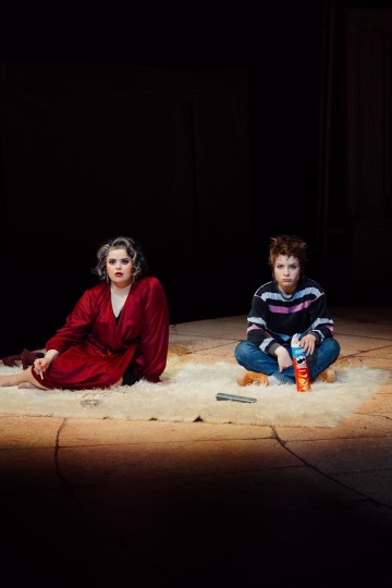 Kaksi ihmistä istuu pyöreällä valkoisella matolla valonheittimen alla. Toisella on punainen kaapu ja meikki, ja hän katsoo eteenpäin; toisella on raidallinen pusero ja farkut, ja hänellä on kädessään Pringles-purkki. Molemmat vaikuttavat vakavilta ja istuvat lavalla.