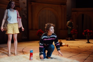 Raitapaitainen henkilö polvistuu valkoiselle matolle dramaattisessa näyttämöympäristössä, ja lähellä on kermavaahtopurkki. Vieressä seisoo toinen henkilö valkoisessa toppatakissa ja keltaisissa shortseissa; taustalla näkyy kaksi muuta.