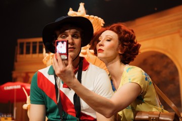 Punatukkainen nainen keltaisessa kukkamekossa ottaa selfien miehen kanssa, jolla on aurinkohattu ja monivärinen paita. He poseeraavat koristeellisen taustan edessä lämpimässä valaistuksessa.