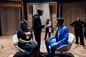 Kaksi ihmistä istuu vastakkain valkoisilla tuoleilla lavalla, toinen sinisessä puvussa ja toinen t-paidassa. Kolme muuta seisoo lähellä, yksi meikkaa, ja taustalla on verhot ja näyttämövalot.