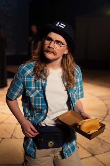 Pitkät hiukset, silmälasit ja viikset, ruudullinen paita, Suomen lippu, valkoinen t-paita, sininen vyölaukku ja tarjotin, jossa on hodari ja ranskalaisia. Tausta on hämärästi valaistu.