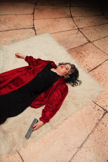 Punaiseen takkiin ja mustaan mekkoon pukeutunut henkilö makaa valkoisella matolla, silmät kiinni, kaukosäädin kädessä, kaakeloidulla lattialla.