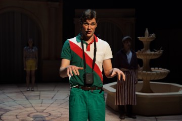 Keskellä näyttämöä seisoo vihreäpunaiseen asuun pukeutunut nuori mies, jolla on kamera kaulassaan. Hän elehtii ilmeikkäästi, kun kaksi muuta ihmistä ja kivilähde näkyvät hämärässä taustalla.