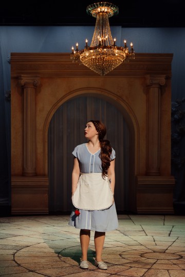 Siniseen mekkoon ja valkoiseen esiliinaan pukeutunut nainen seisoo lavalla, jossa on kivilattia, ja katsoo ylös hehkuvaan kattokruunuun. Taustalla on suuria pylväitä ja kaareva oviaukko, jotka luovat suurenmoisen, tyylikkään ympäristön.